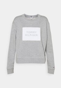 Gray sweatshirt with a white rectangular patch featuring "TOMMY HILFIGER" in embossed text. Ribbed cuffs and crew neckline. Fabric texture is soft.