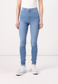 Svijetloplave skinny traperice visokog struka s diskretnim šavovima, bočnim džepovima i glatkom traper teksturom, uparene s bijelim visoko-top adidasicama.