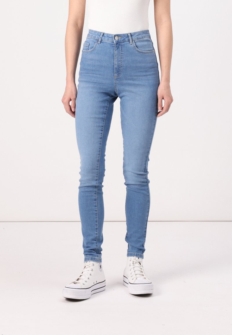 Svijetloplave skinny traperice visokog struka s diskretnim šavovima, bočnim džepovima i glatkom traper teksturom, uparene s bijelim visoko-top adidasicama.