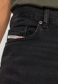 Jeans de mezclilla negra con un bolsillo frontal que exhibe una etiqueta roja y blanca. Las características incluyen un botón de metal y una tela texturizada.