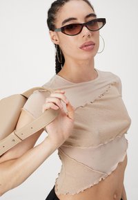 Šis gaiši brūns īsais tops ar viļņainiem ruffles akcentiem izgatavots no mīksta auduma. Modelis tur nedaudz rozā siksniņu somiņu, valkājot taisnstūra saulesbrilles un riņķa auskarus.