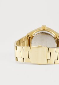 Guldmetallænke til ur med lås, indgraveret med "GUESS", fastgjort til et gyldent armbåndsur med synlig krone og bagside.