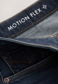 Primo piano della cintura di jeans blu scuro con etichetta bianca "MOTION FLEX" e dettagli di cuciture marroni.