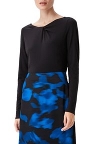 Langärmliges Oberteil in Schwarz mit Rundhalsausschnitt und Drehdetails, kombiniert mit einem knielangen Rock mit einem blau-schwarzen abstrakten Muster.