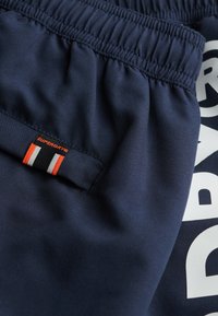 Shorts de sport bleu marine avec une ceinture élastique, comportant un logo blanc et une étiquette en tissu avec des rayures rouges, blanches et orange.