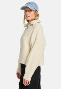 Cremfarbener, strukturiertes Sweatshirt mit Kragen und weiten Ärmeln, kombiniert mit einer hellblauen Jeansmütze. Schwarze Hosen sind darunter sichtbar.