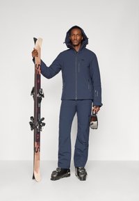 Giacca da sci blu marina con cappuccio e cerniere, pantaloni coordinati e scarponi da sci neri. In mano sci con attacchi e occhiali da sci.