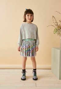 Felpa grigia corta, gonna svasata con paillettes arcobaleno e stivali neri con lacci e accenti iridescenti. Sfondo semplice e colori neutri.