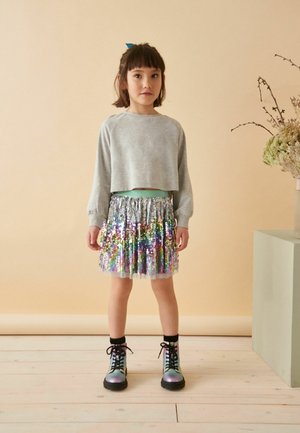 Grauer, kurzer Pullover, regenbogenfarbiges Pailletten-Minirock und schwarze Schnürstiefel mit schillernden Akzenten. Schlichter Hintergrund und neutrale Farben.