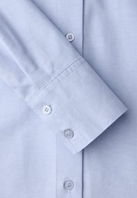 Vaaleansininen kauluspaita, jossa on sileä kangas, napillinen nappilista ja neljän napin sekä ommeltujen reunoiden kalvosimet.