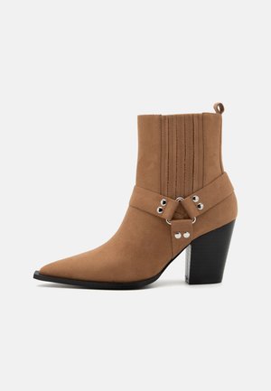 Stivaletto alla caviglia in suede marrone con punta a punta, texture a costine sui lati, cinturino regolabile con dettagli argento e tacco chunky nero.