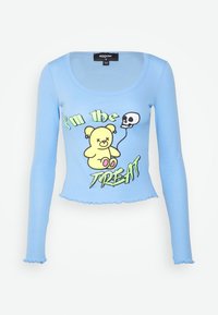 Svetle modrý dlouhý rukáv crop top s kulatým výstřihem, zdobený grafickým motivem karikatury medvěda, lebkovým balónem a textem "Jsem PŘÍSNĚ DOBRÝ" v zelené barvě.