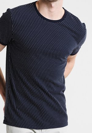 Mann trägt ein eng anliegendes marineblaues T-Shirt mit kleinen weißen Punkten, steht vor einem schlichten hellen Hintergrund.