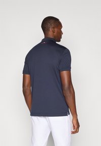 Polo de cor azul marinho com colarinho, mangas curtas e rachas laterais; apresenta um logótipo vermelho nas costas, perto da gola; tecido liso e leve.