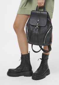 Czarny skórzany plecak z teksturowanym wykończeniem, z klapą na zamek, srebrnymi akcentami i dwiema kieszeniami na zamek. Czarne, masywne buty bojowe.