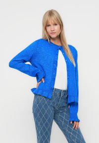 Cardigan bleu vif avec un col rond, boutonnière et ourlet festonné ; porté sur un haut blanc et associé à un jean en denim orné.
