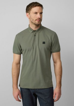 Olivgrünes Poloshirt aus strukturiertem Baumwollstoff mit einer Knopfleiste mit zwei Knöpfen und einem kleinen schwarzen Logo-Patch auf der Brust. Kurze Ärmel.