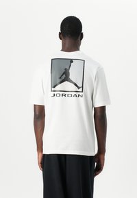 Hvid bomulds t-shirt med en stor sort grafik af en springende figur og "JORDAN" tekst på ryggen. Korte ærmer og afslappet pasform.