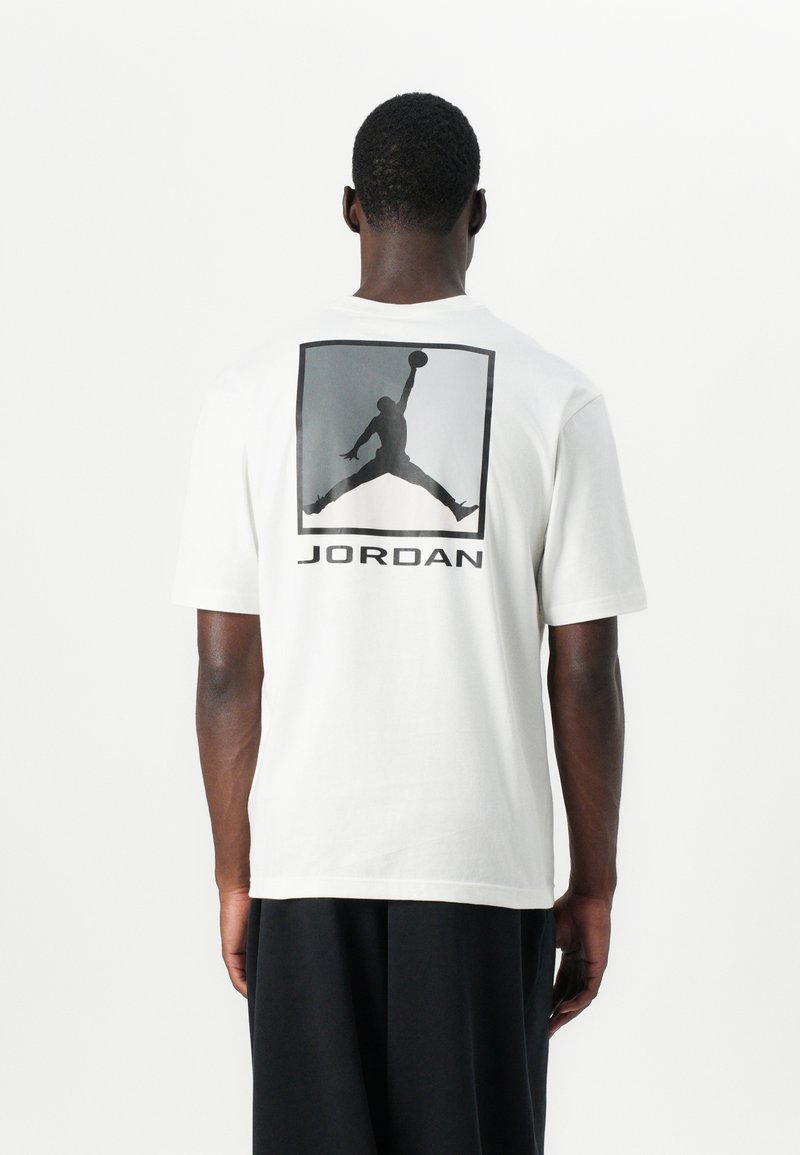 Hvid bomulds t-shirt med en stor sort grafik af en springende figur og "JORDAN" tekst på ryggen. Korte ærmer og afslappet pasform.