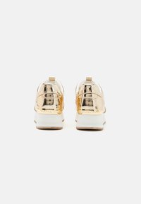 Adidași metalici aurii cu vârf rotund, talpă albă și detaliu texturat la spate cu branding. Mâner la spate pentru o purtare ușoară.