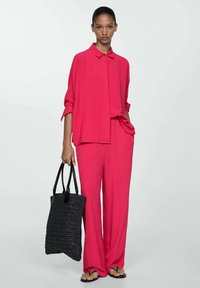 Fuchsia tvådelad set med en knapp-up skjorta och vida byxor, tillverkad av lättviktig tyg. Modellen bär en svart vävd strandväska.