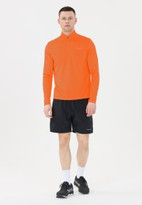 Kirpeä oranssi pitkähihainen urheilupaita, jossa on puolivetoketju, yhdistettynä mustiin SHORTSeihin. Rakenteellinen kangas, kevyt muotoilu ja erottuvat asusteet.
