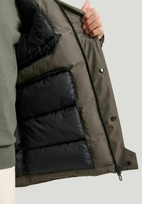 Puffjacka med fodrad insida; ytterdel i mörk olivgrön, innerside i svart, med dragkedja och tryckknappar för stängning.