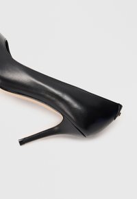 Chaussure à talon haut en cuir noir avec un bout pointu et des côtés ouverts. Dotée d'un talon aiguille fin et d'une texture lisse.