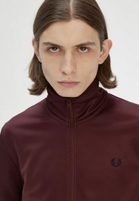 Giacca bordeaux con zip e colletto alto, con logo ricamato sul lato sinistro del petto. Tessuto liscio e opaco, design aderente e silhouette slim.
