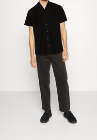 Chemise noire à manches courtes avec un devant à boutons et une texture douce, associée à un pantalon gris foncé et des chaussures noires.