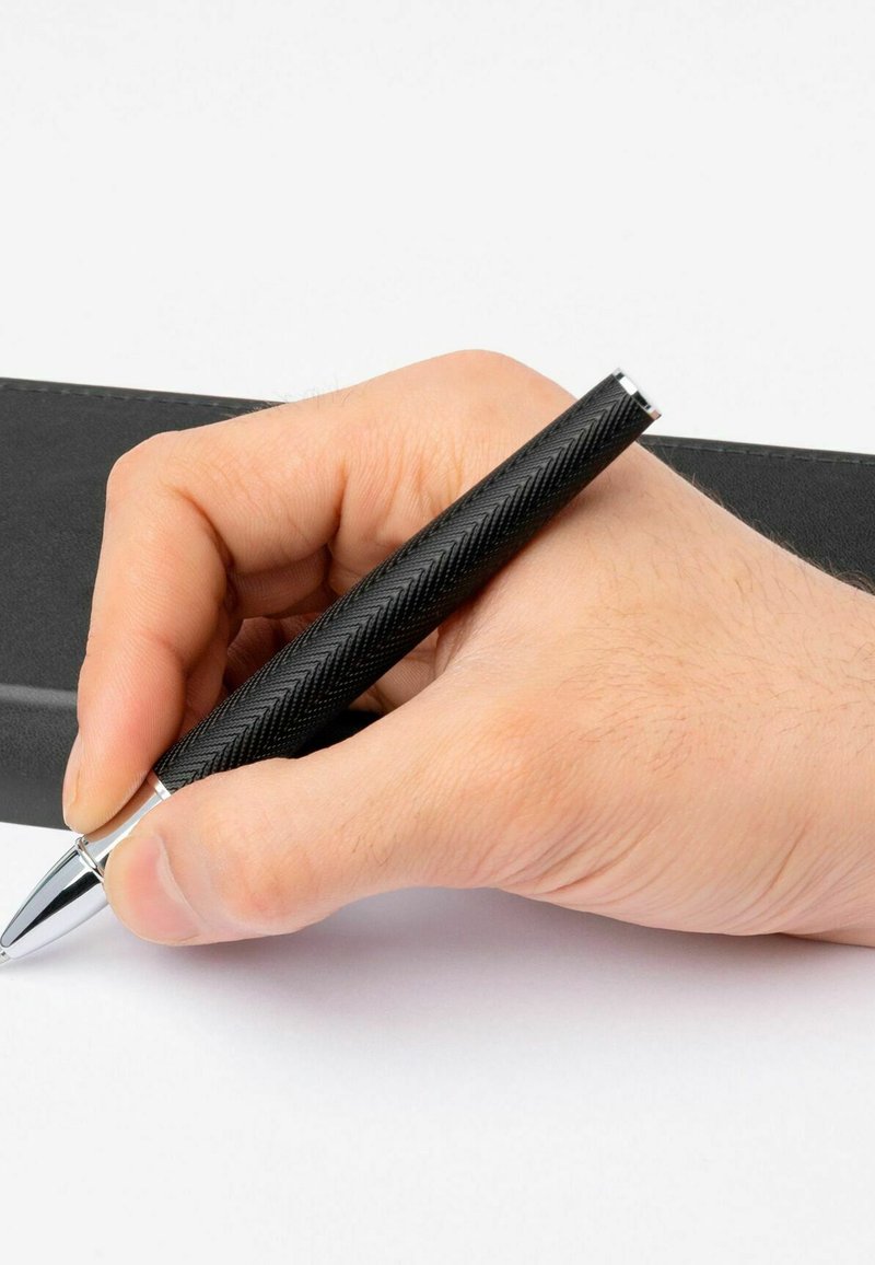 Bolígrafo de textura negra con patrón de espiga, detalles en cromo, sostenido en una mano sobre papel blanco, junto a una carpeta negra.
