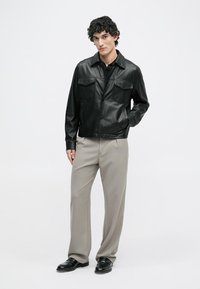 Μαύρο δερμάτινο μπουφάν με δύο τσέπες στο στήθος, κλείσιμο με κουμπί και μυτερό γιακά. Φοριέται με ανοιχτό γκρίζες καρό παντελόνες και μαύρα παπούτσια.