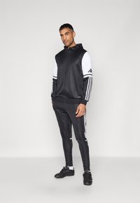 Tuta sportiva nera con accenti bianchi, composta da una giacca con cappuccio e zip e pantaloni affusolati. Logo con tre strisce sulle maniche e sui lati.