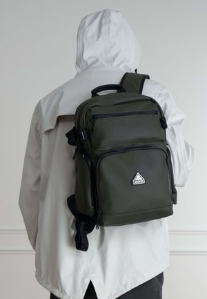 Zaino verde scuro realizzato in materiale liscio, con diverse tasche con zip, tracolla e una piccola toppa a forma di triangolo.