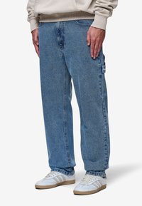 Jeans de mezclilla azul claro con un corte relajado, que cuentan con un bolsillo lateral y una sutil textura descolorida. Combinados con zapatillas deportivas blancas y una sudadera beige.