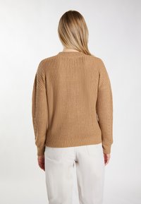 Beżowy sweter z dzianiny o teksturowanym wzorze, z prążkowanymi mankietami i dołem, w luźnym kroju. Ubrany z jasnymi spodniami, widok z tyłu.