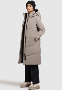 Long, padded coat in light beige, featuring a hood, zip and button front closure, side pockets, and a straight fit. Worn over dark pants and chunky shoes.