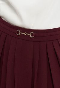 Ζώνη από βυσσινί πλισέ φούστα με λεπτή ταιριαστή ζώνη που διαθέτει μια μικρή χρυσή αγκράφα σε στυλ χαλινού αλόγου.