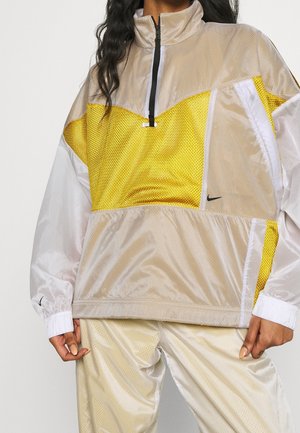 Personne portant une veste de sport Nike beige et jaune avec une fermeture éclair à l'avant et un pantalon de survêtement beige assorti.