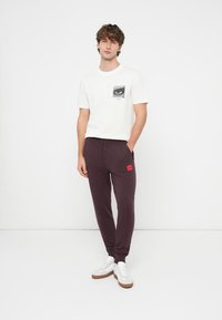 Men's white t-shirt with a black-and-white eye graphic and red logo on the chest, paired with dark burgundy sweatpants featuring a red patch.
