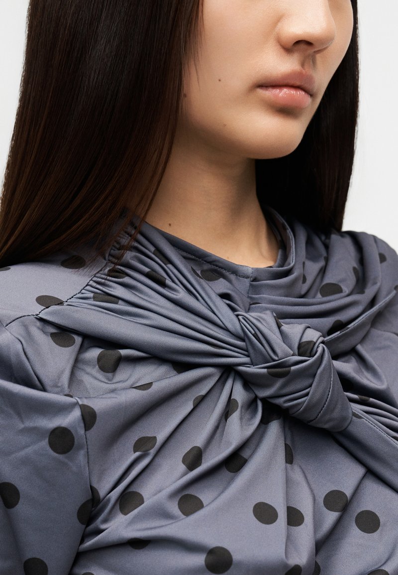 Haut en tissu gris avec un détail de nœud au niveau de l'encolure et un motif de points noirs. Le matériau semble lisse avec un léger éclat.