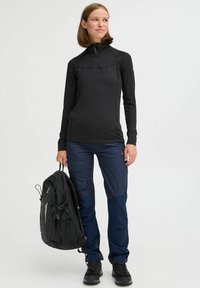 Zwarte lange mouwen top met een halve rits, marineblauwe cargobroek met zakken en zwarte wandelschoenen. Een zwarte rugzak wordt in één hand vastgehouden.