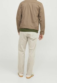 Chaqueta de lona marrón sobre una camisa verde, combinada con jeans de color claro y zapatillas. Costuras visibles y detalles del bolsillo trasero en los jeans.