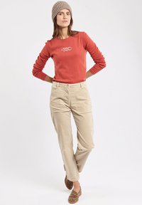 Donna che indossa un cappello di maglia beige, una maglietta a maniche lunghe color ruggine con logo, pantaloni beige e mocassini con stampa leopardata, in piedi con le mani sui fianchi.