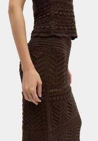 Vista lateral de una persona que lleva un top de crochet marrón oscuro y una falda a juego con patrones geométricos intrincados.