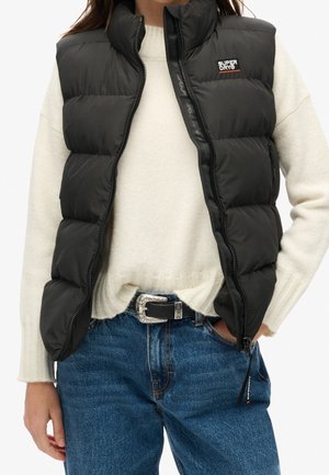 Vrouw draagt een zwart doorgestikt mouwloos vest met Superdry-logo, off-white trui, blauwe spijkerbroek en een zwarte riem met zilveren gesp.