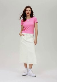Haut rose à manches courtes avec un détail en nœud, associé à une jupe midi en denim blanc. Le mannequin porte des baskets blanches et a de longs cheveux ondulés.
