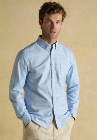 Muž s kučeravými vlasmi, oblečený v svetlomodrej kockovanej košeli s gombíkmi a béžových nohaviciach, stojaci s rukami v vreckách na neutrálnom pozadí.