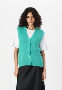 Turquoise knitted vest with a V-neck, five buttons, and ribbed texture, layered over a white T-shirt, paired with a black skirt.