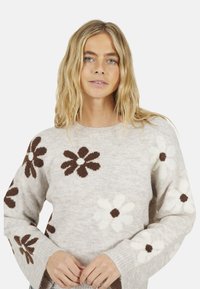 Femme blonde portant un pull beige avec de grands motifs floraux bruns et blancs, se tenant devant un fond uni.
